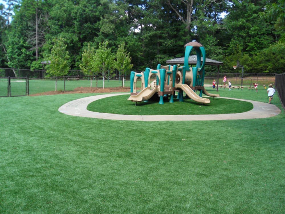 Park playground with slides and climbing structures on artificial turf surrounded by trees