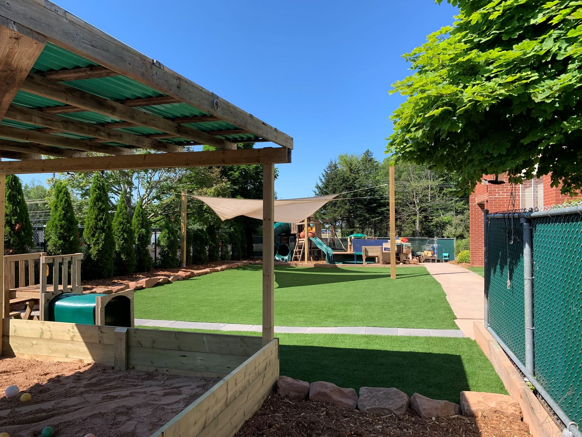 Daycare playground with covered sandbox, play structure, and shade sails on artificial grass